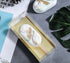 A clear PVC box contains a decorative cakesickle, resembling a popsicle. The cakesickle is predominantly white with green and gold accents, featuring a design that includes two white feather shapes and gold leaf detailing. The box has a gold-colored base and a wooden stick protruding from the bottom of the cakesickle. In the background, there are some leaves and a textured surface, along with partially visible printed materials.