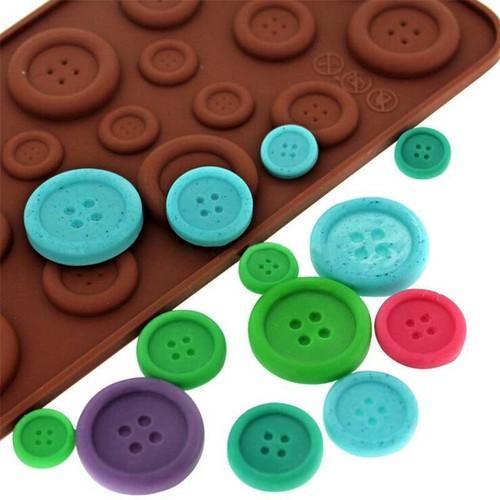 A silicone mould tray shaped like buttons in various sizes and colors. The mould is brown, and there are several colorful button-shaped pieces scattered around it, including blue, green, pink, and purple buttons, each featuring four holes.