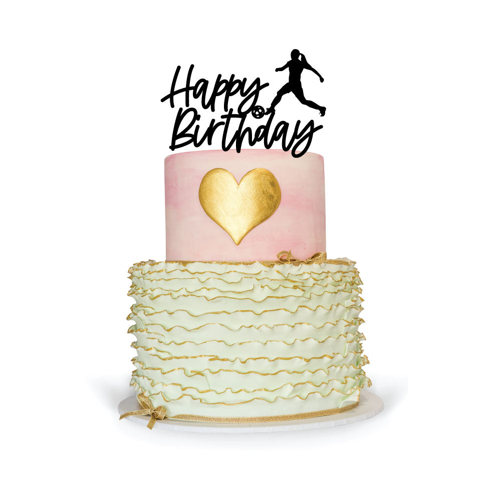 A two-tier birthday cake featuring a pink top tier with a gold heart design, and a bottom tier decorated with white ruffles and gold accents. Atop the cake is a black custom cake topper that reads &quot;Happy Birthday&quot; with a silhouette of a soccer player in action. The cake is presented on a circular white plate against a white background.