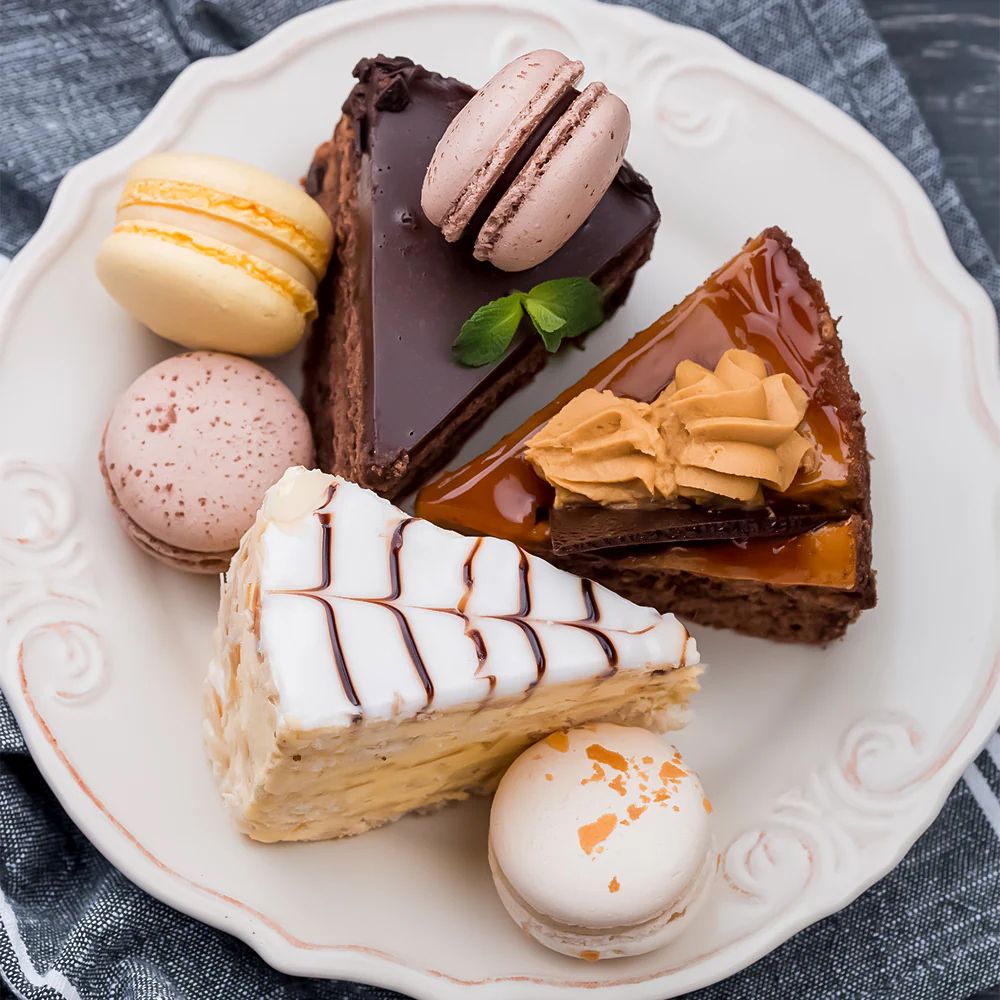 A plate with three cake slices—chocolate, caramel, and vanilla—each topped with macarons in pastel shades, creating a delightful and elegant dessert display.
