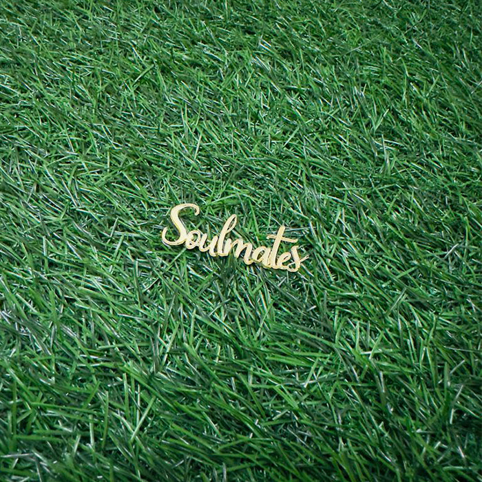 A wooden cake topper that reads "Soulmates" in an elegant cursive font, placed on a textured green grass background.
