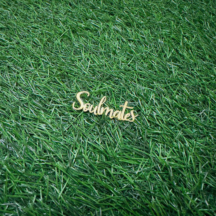 A wooden cake topper that reads &quot;Soulmates&quot; in an elegant cursive font, placed on a textured green grass background.