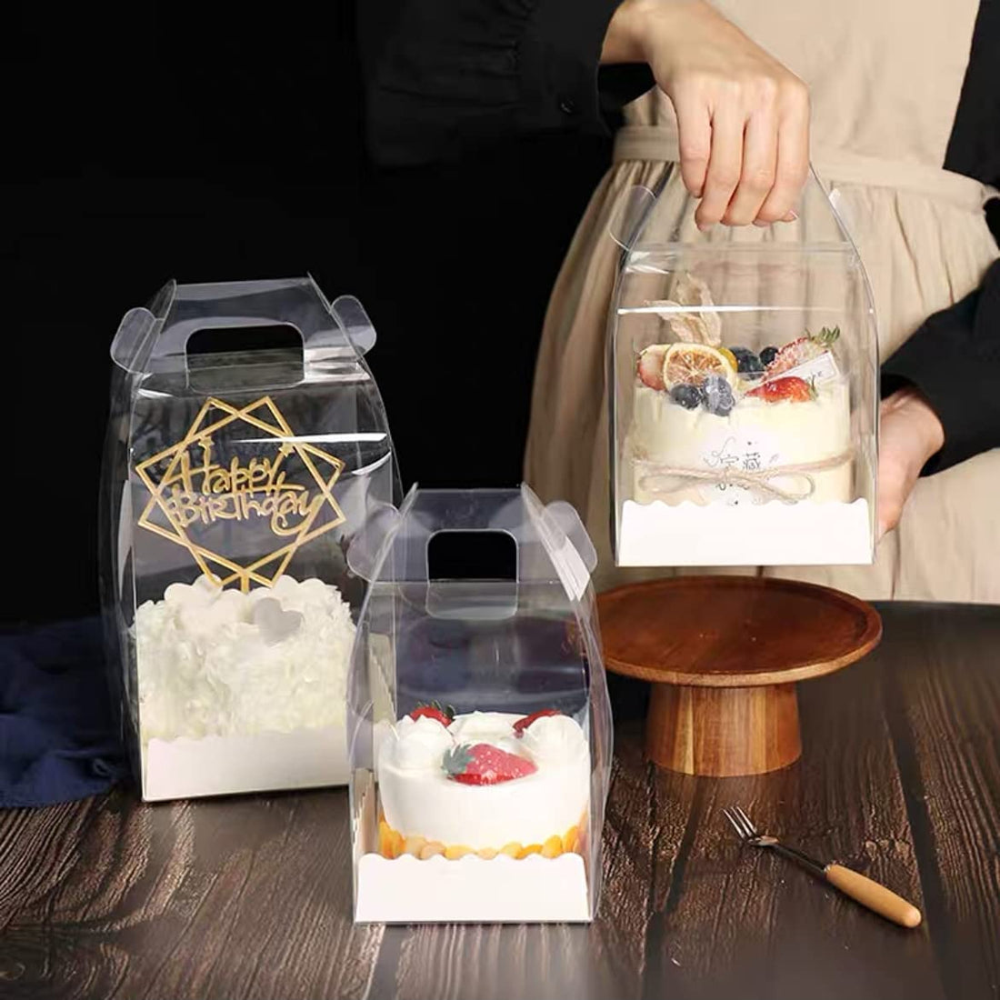 The image depicts three square, transparent PVC cake boxes displayed on a wooden table with a round wooden cake stand in the background. Each box contains a different cake. The box on the left features a cake topped with decorative elements and a "Happy Birthday" sign. The middle box contains a cake adorned with strawberries and cream. The right box holds a cake decorated with assorted fruits, including blueberries and citrus slices. A small fork can be seen next to the cake stand.