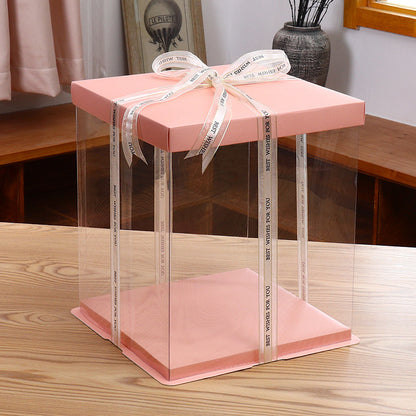A square cake box with dimensions 14 x 14 x 14 inches, featuring a soft pink lid and clear sides. The box is tied with a decorative ribbon that has the text "BEST WISHES FOR YOU" printed on it. It is placed on a wooden table, and there is a window in the background.