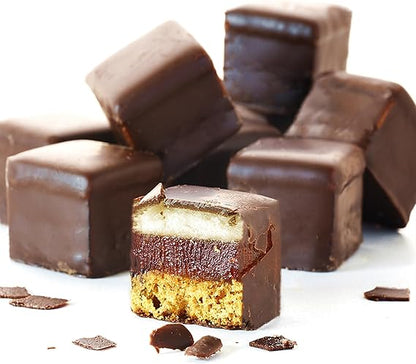 A close-up image of square-shaped chocolate confections is displayed. Several dark chocolate-covered cubes are stacked in the background, while one cube is cut in half to reveal its layered interior. The exposed section shows three distinct layers: a chocolate top layer, a fruit or jelly middle layer, and a crumbly base layer, all resting on a clean white surface. Small chocolate shavings are scattered around.