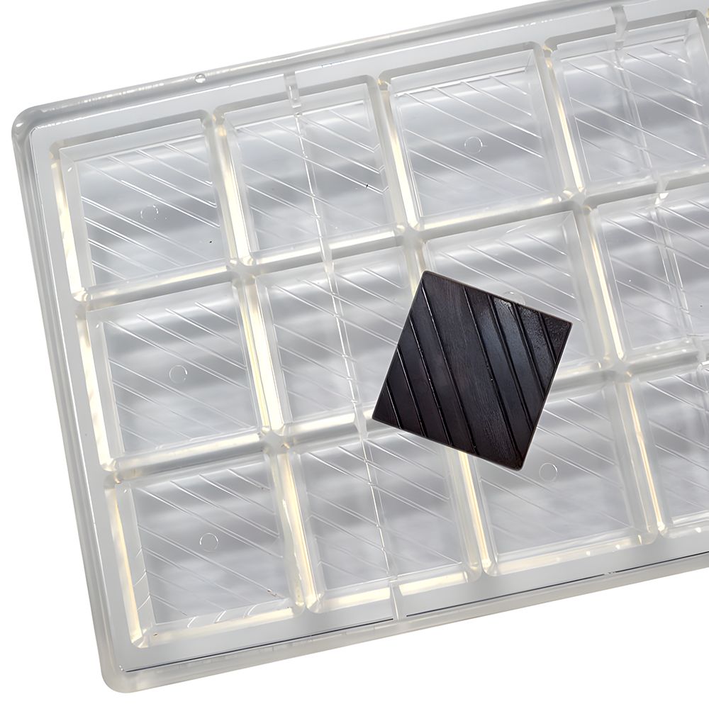 A transparent plastic tray with 16 rectangular compartments, one empty. A single dark chocolate square is placed at an angle on the tray, suggesting indulgence.