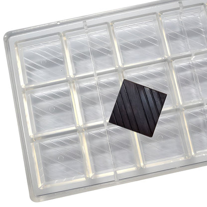 A transparent plastic tray with 16 rectangular compartments, one empty. A single dark chocolate square is placed at an angle on the tray, suggesting indulgence.