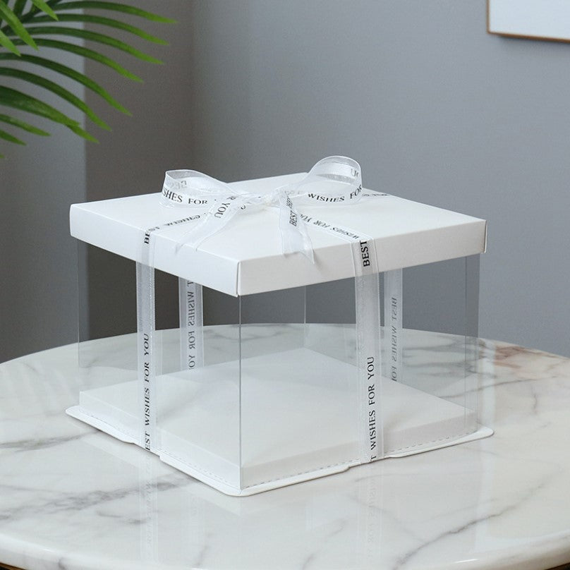 A square cake box made of white PVC with a transparent front and sides. It features a white lid and is adorned with a ribbon tied in a bow, printed with the text &quot;BEST WISHES FOR YOU&quot; in black. The box sits on a marble surface with a plant visible in the background.