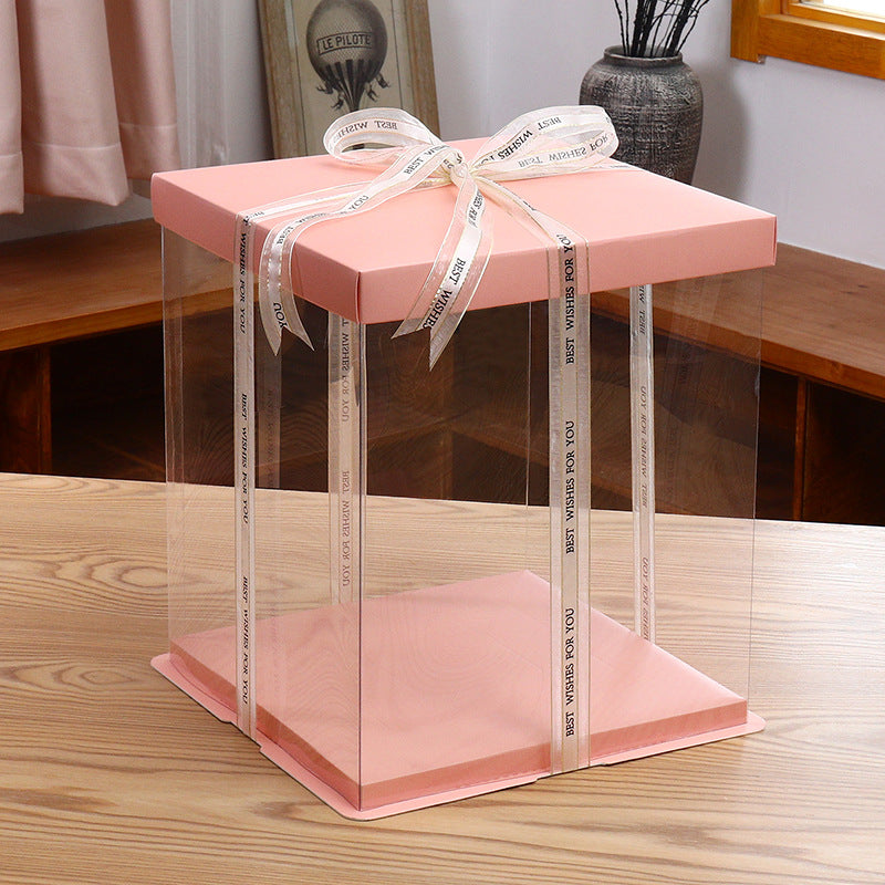 A square cake box with a pink lid and clear sides, measuring 10 x 10 inches and 9.8 inches high. It features a decorative ribbon tied in a bow at the top, with the text &quot;BEST WISHES FOR YOU&quot; printed on it. The box is placed on a wooden surface with a light-colored finish, and there are soft furnishings and decor in the background.