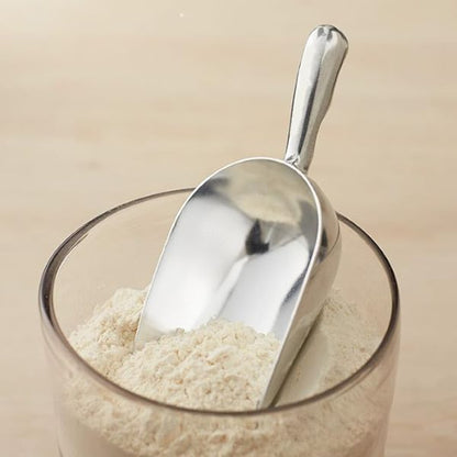 A stainless steel scoop is resting on top of a glass container filled with flour. The scoop has a smooth, shiny surface and a curved shape, partially submerged in the fine white powder. The background is a light wooden surface.