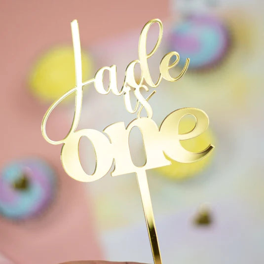 A gold acrylic cake topper features the text &quot;Jade is one.&quot; The topper has an elegant, cursive font for the word &quot;Jade&quot; and a bold font for &quot;is one.&quot; The background is a soft pink with blurred pastel-colored cupcakes visible.