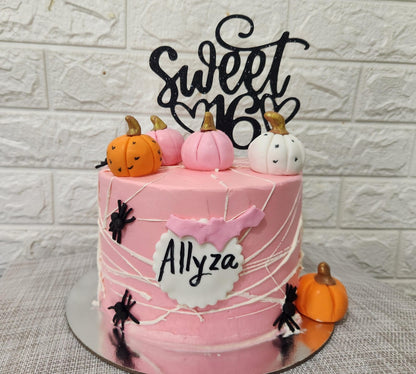 A tiered pink birthday cake adorned with a black cardstock topper that says "Sweet 16." The cake is decorated with small fondant pumpkins in orange and white, and has black spider decorations on the sides. A pink flag with the name "Allyza" is attached to the front of the cake. The background features a light-colored, textured wall.