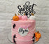 A tiered pink birthday cake adorned with a black cardstock topper that says "Sweet 16." The cake is decorated with small fondant pumpkins in orange and white, and has black spider decorations on the sides. A pink flag with the name "Allyza" is attached to the front of the cake. The background features a light-colored, textured wall.