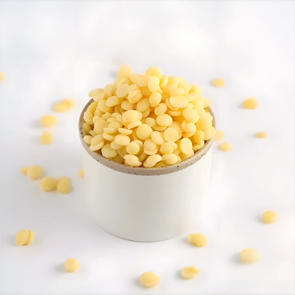 A white ceramic cup filled with small, yellow beeswax pellets sits on a white surface. Some pellets are scattered around, creating a clean, minimalistic look.