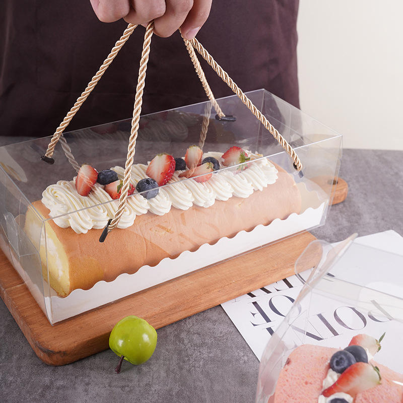 A person is holding a transparent box with handles, containing a Swiss roll dessert decorated with whipped cream and topped with sliced strawberries and blueberries. The box is placed on a wooden cutting board, and there&