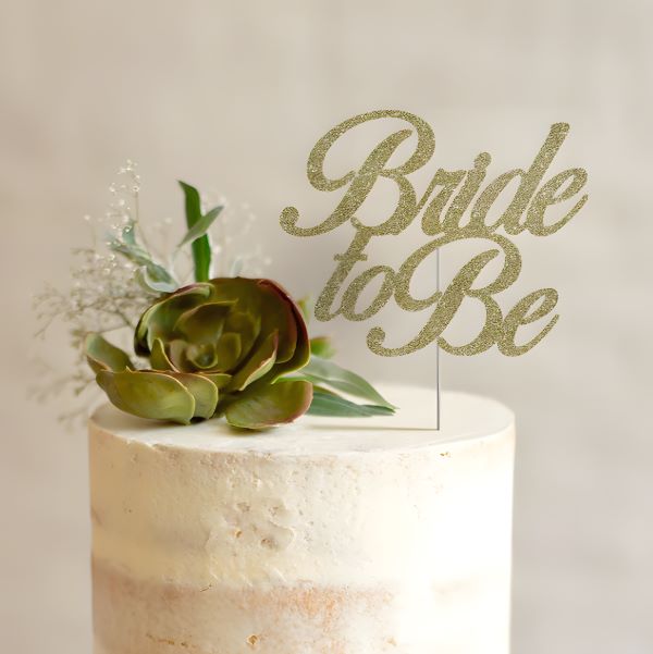 A cream-colored, frosted cake topped with a gold glitter cardstock cake topper that reads &quot;Bride to Be.&quot; The topper is decorated with a succulent and green leaves, along with small white flowers. The background is softly blurred.