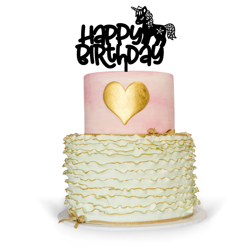 A two-tier birthday cake with a unicorn theme. The top tier is pink with a golden heart-shaped decoration, while the bottom tier features green icing with ruffled texture and gold accents. A black cake topper reads &quot;Happy Birthday&quot; accompanied by a unicorn silhouette. The cake is placed on a white circular base.