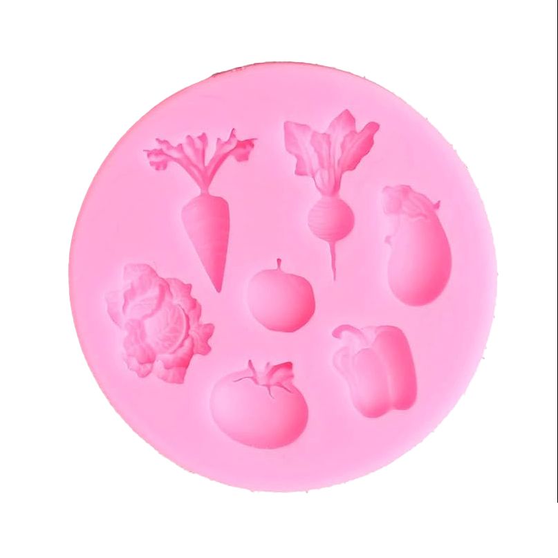 A round pink silicone mold depicts seven recessed vegetable shapes: carrot, radish, eggplant, cauliflower, apple, tomato, and bell pepper.