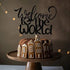 An image depicts a chocolate cake topped with a customized cardstock topper that reads "Welcome to the World" in an elegant, cursive font. The cake is garnished with gingerbread houses around its base, and it is displayed on a wooden cake stand. The background is softly illuminated with string lights, creating a warm atmosphere.