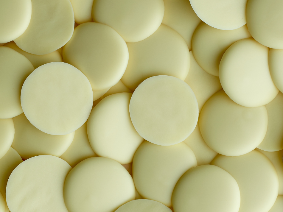 A close-up view of multiple round, flat coins of white chocolate, arranged closely together. The coins are light yellowish-white in color with a smooth surface, and they vary slightly in size and orientation, creating a visually appealing pattern.