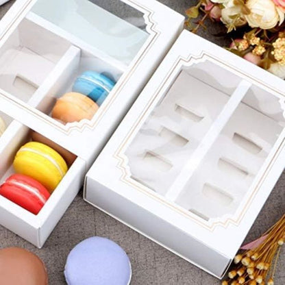 The image features a product called "window macaron box 10 portions." There are two white boxes displayed, each with a clear window. One box contains four brightly colored macarons: red, yellow, orange, and blue. The other box is empty, showing six compartments designed for macarons. Soft pastel-colored macarons, like a lavender one, are also visible on the surface beside the boxes. The background includes decorative dried flowers, enhancing the overall presentation.