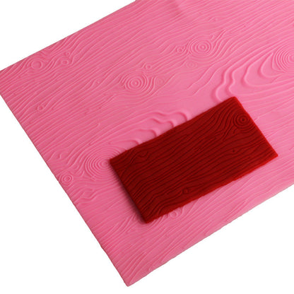 An impression silicone mat is displayed, featuring a textured surface designed to resemble wood grain. The mat is predominantly pink and showcases various wood-like patterns with grooves and knots. A small rectangular section of red silicone, also with wood grain texture, is placed on top of the pink mat. The overall layout emphasizes the mat&