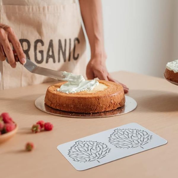 A person wearing an apron that reads "ORGANIC" is spreading cream on top of a round cake placed on a silver cake board. The cake sits on a wooden table with a plate of raspberries nearby. In the foreground, there is a silicone mold featuring decorative leaf patterns. Another cake can be seen in the background, partially obscured. The overall scene is bright and focused on cake decorating.