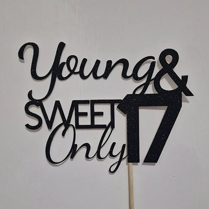 The image features a cake topper made of black glitter cardstock. The design includes the phrases "Young &amp; Sweet" in a flowing script font, with "Only" in a bold, capitalized font. The number "17" is prominently displayed, also in a bold font, indicating a celebration for a 17th birthday. The topper is mounted on a stick for easy placement on a cake, and it is set against a plain background.